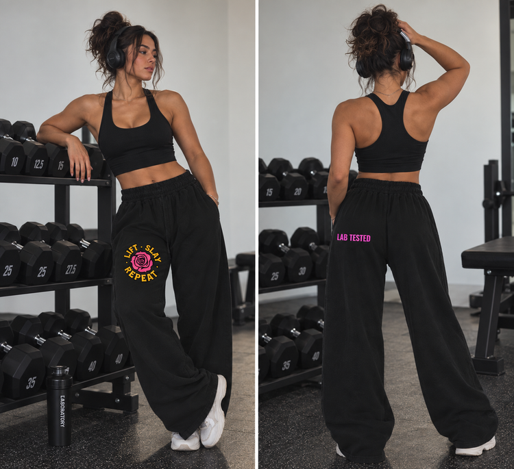 A split view of a woman in a gym wearing black athletic sweatpants with graphic prints. The front view shows a black sports bra and black sweatpants with a pink rose and yellow 'JUST SLAY EVERY' graphic on the left thigh.