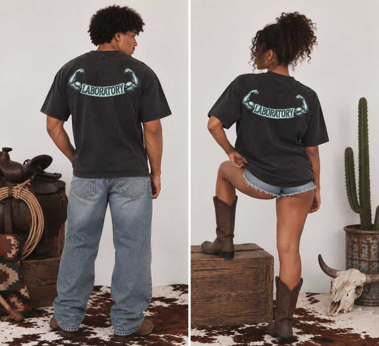 A split-screen lifestyle image showing a man and a woman from the back wearing a dark charcoal gray t-shirt with a western-style graphic that reads 'LABORATORY' with flexing muscles. The man wears light blue jeans and stands next to a rustic saddle, while the woman wears denim shorts and brown cowboy boots, posing on a wooden crate in a western-themed setting.