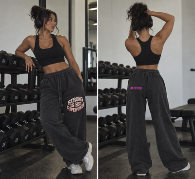 A woman modeling black athletic sweatpants in a gym setting, shown from both the front and back. The front view shows the front of the black sports bra and the back view shows the back of the black sweatpants with pink and white text graphics.