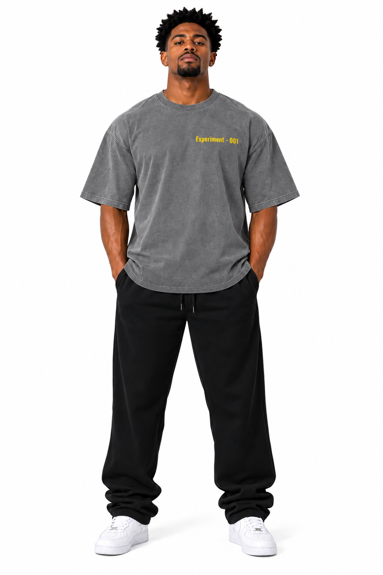 A man modeling a heather gray t-shirt with yellow 'Experiment-001' text on the chest, paired with black sweatpants and white sneakers, against a plain white background.