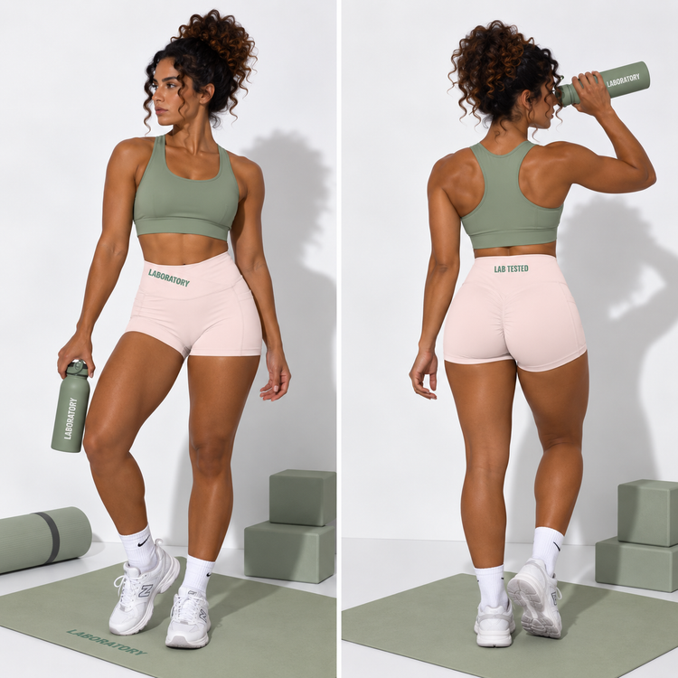 A split front and back view of a woman modeling a sage green sports bra and light pink athletic shorts with 'LAB ORDER' printed on them, standing on a yoga mat with green blocks in a studio setting.