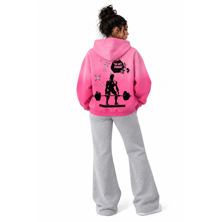 A model shown from the back wearing a bright pink hoodie with a black graphic print on the back and light gray sweatpants, set against a plain white background.