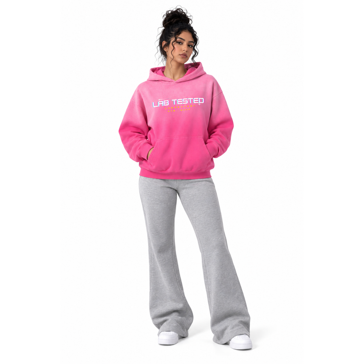 A woman modeling a bright pink hoodie with 'LAB TESTED' printed in white across the chest, paired with light gray sweatpants and white sneakers, set against a plain white background.